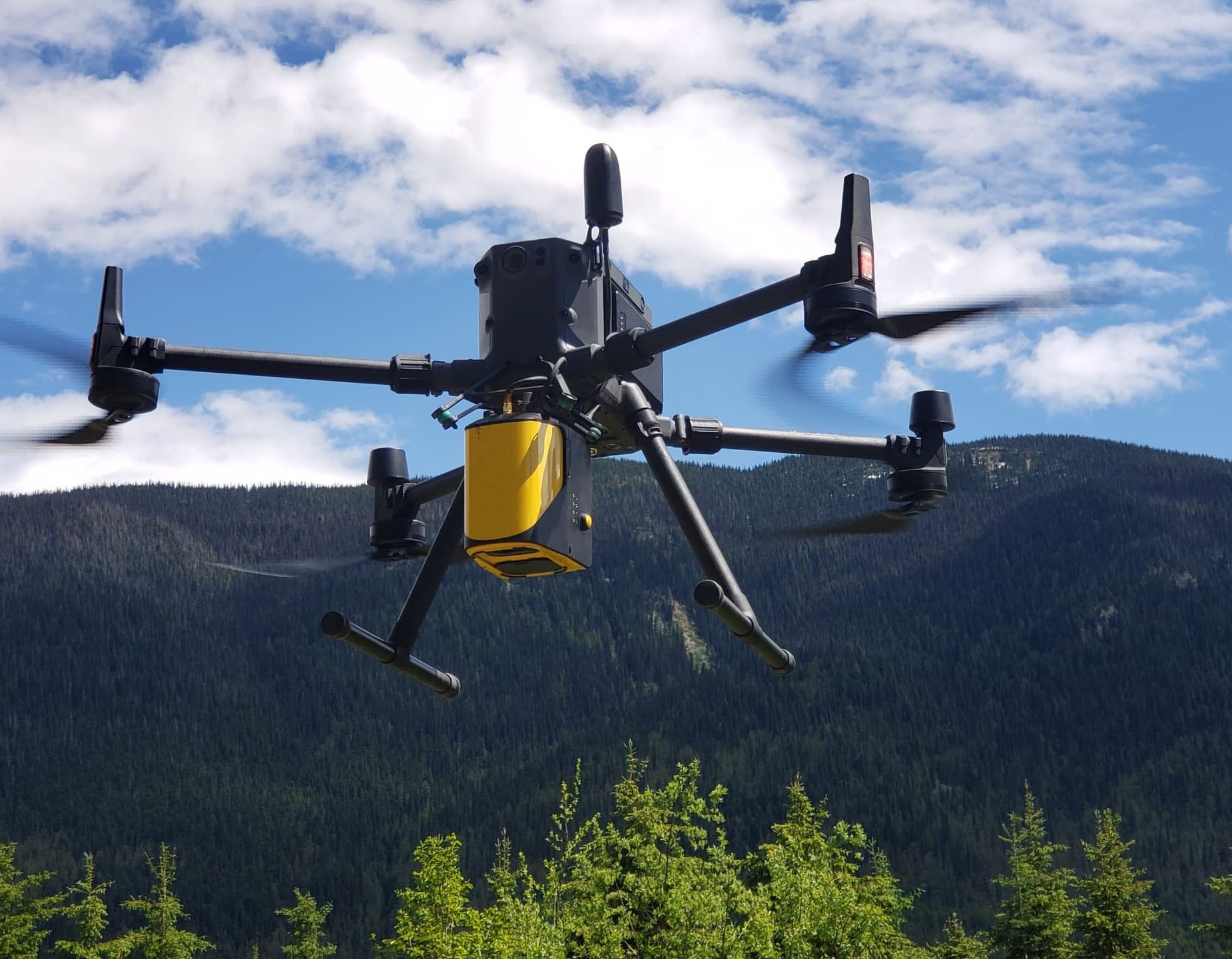 A black and yellow drone flying under cloudy blue skies over a forested hillside for strategic natural resource consultancy